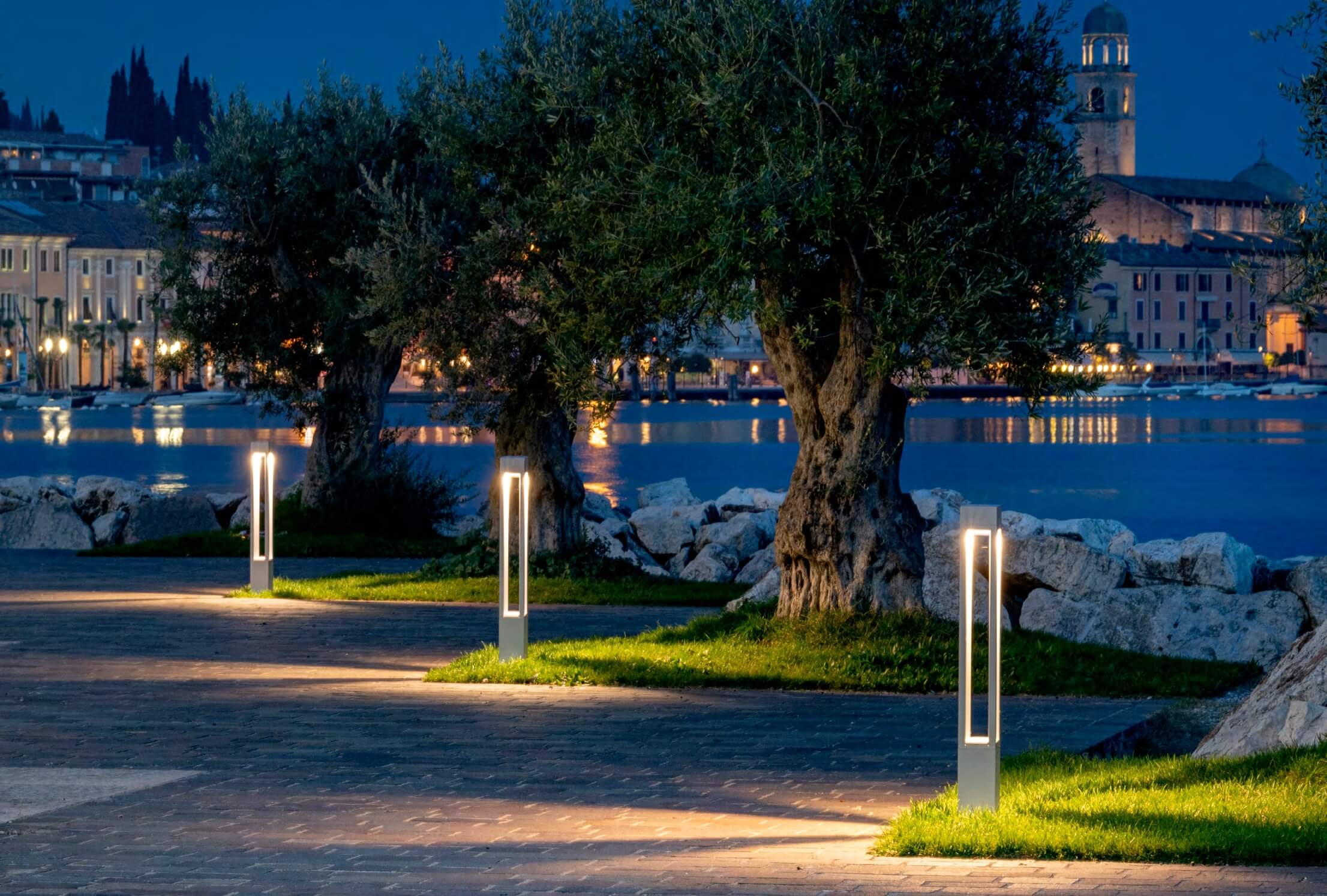 Platek Pollerleuchten beleuchten eine Uferpromenade zwischen Olivenbäumen bei Dämmerung